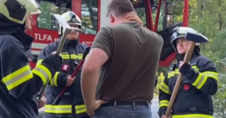 A man stands between two firefighters, holding his back. Both firefighters hold axes, and one wears a helmet. A fire truck is in the background.