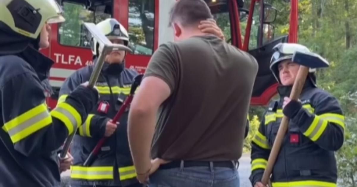 A man stands between two firefighters, holding his back. Both firefighters hold axes, and one wears a helmet. A fire truck is in the background.