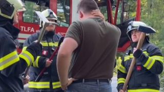 A man stands between two firefighters, holding his back. Both firefighters hold axes, and one wears a helmet. A fire truck is in the background.