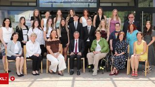 A group of individuals, including men and women, stand and sit in front of a building for a photo. Some wear formal attire, glasses, and accessories. One man is in a wheelchair.