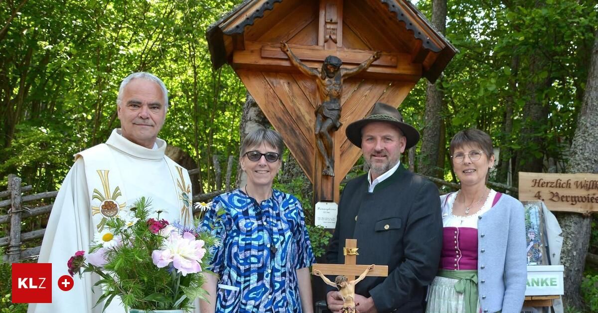 Four people stand in front of a wooden shrine, holding flowers and a cross. A priest, two women, and a man are posing for a photo.