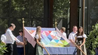 A group of people stand on a stage holding a large quilt. They are in front of a crowd and are dressed in traditional clothing. Trees and a building are visible in the background.