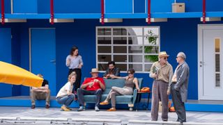 A group of people on a stage with a blue background, some sitting on a blue couch, one standing and holding a book.