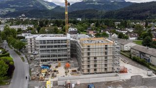Aerial view of a multistory building under construction in a town with mountains in the background. Cranes and scaffolding surround the building.