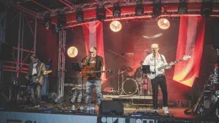 A band performs on a stage with a red backdrop. The lead singer sings into a microphone. Another band member plays a guitar and another plays a bass guitar. The stage is equipped with speakers, a drum set, and spotlights.
