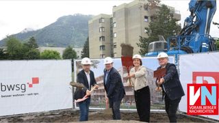 Four people in white hard hats and suits stand in a dirt field, holding shovels, as if breaking ground for a new building project.