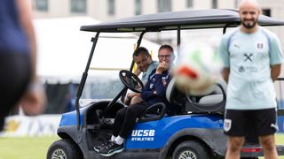 Two men ride a blue golf cart with UCSB Athletics written on the side. One man holds a ball.