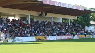 A soccer stadium with a large crowd of spectators, including families and children. The stadium has banners with logos and the name ASV Siegendorp on the roof.