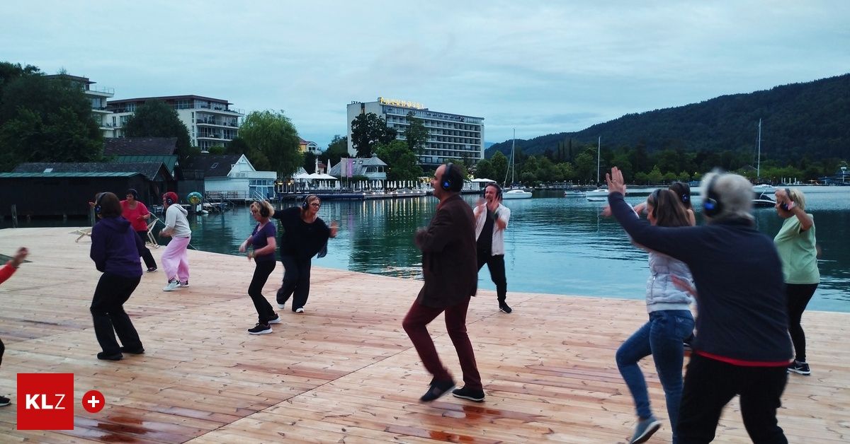 A group of people wearing headphones dance on a wooden dock by the water. Buildings and trees are in the background.