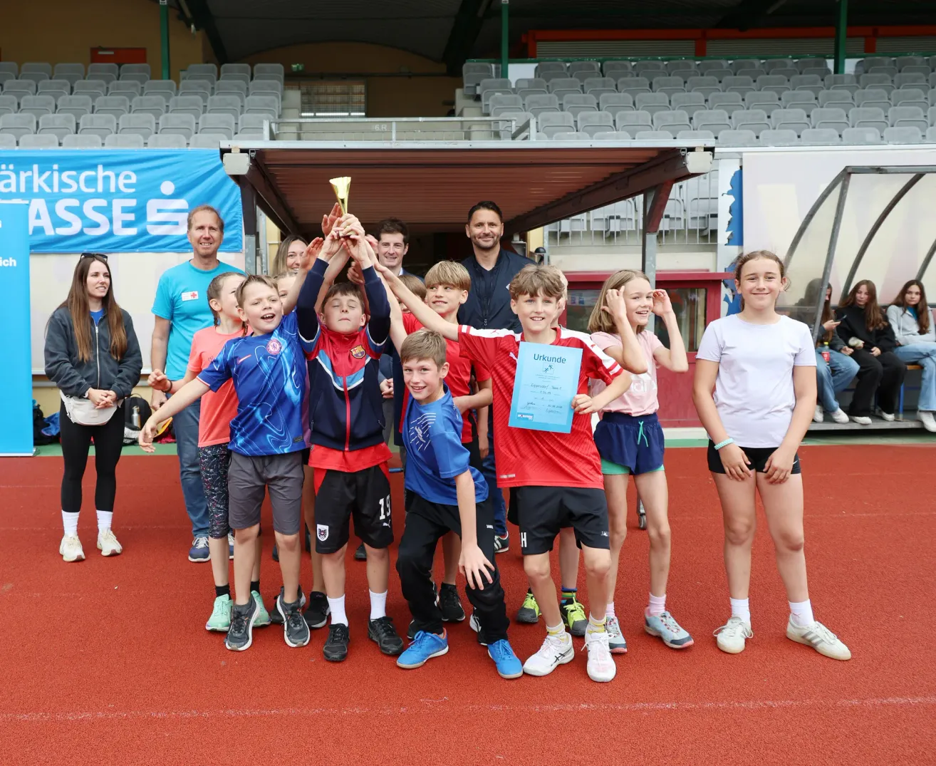 Eine Gruppe von Kindern und Erwachsenen posiert mit Trophäen und Urkunden auf einer roten Laufbahn. Die Erwachsenen stehen im Hintergrund. Die Kinder lächeln und heben ihre Hände. Ein leeres Stadion ist im Hintergrund sichtbar.