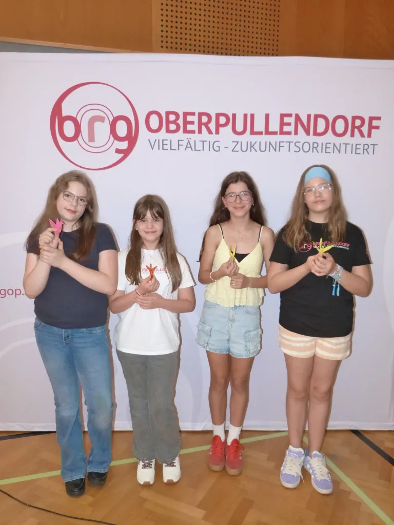Four girls stand in front of a white banner with the text 'Oberpullendorf'. They are smiling and holding paper cranes. They seem to be at an event.