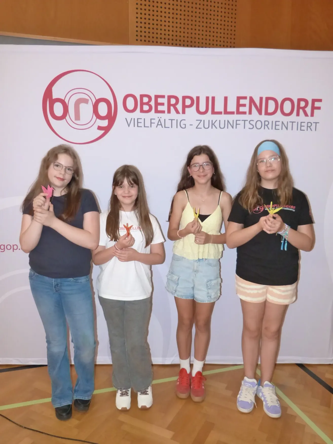 Four girls stand in front of a white banner with the text 'Oberpullendorf'. They are smiling and holding paper cranes. They seem to be at an event.