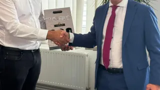 Two men in suits, one holding a gift box, are shaking hands inside an office. Behind them is a white radiator, and a potted plant.