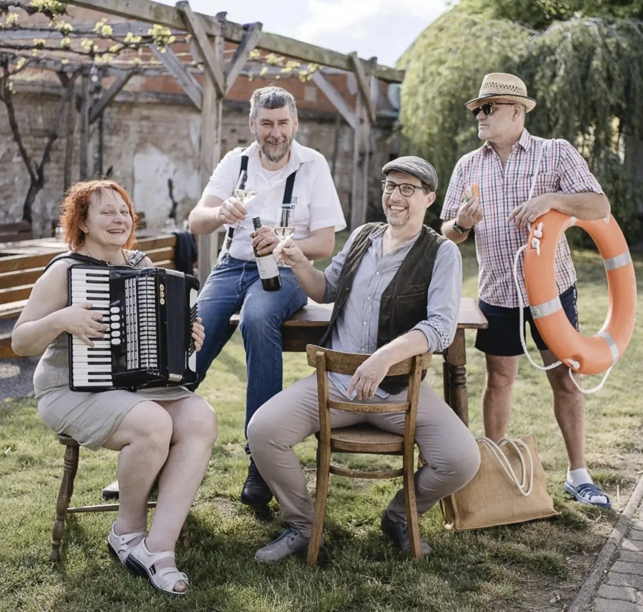Four adults sitting on chairs and benches outdoors, two holding wine glasses, one playing an accordion, one holding a bottle, and a lifesaver hanging on the side.