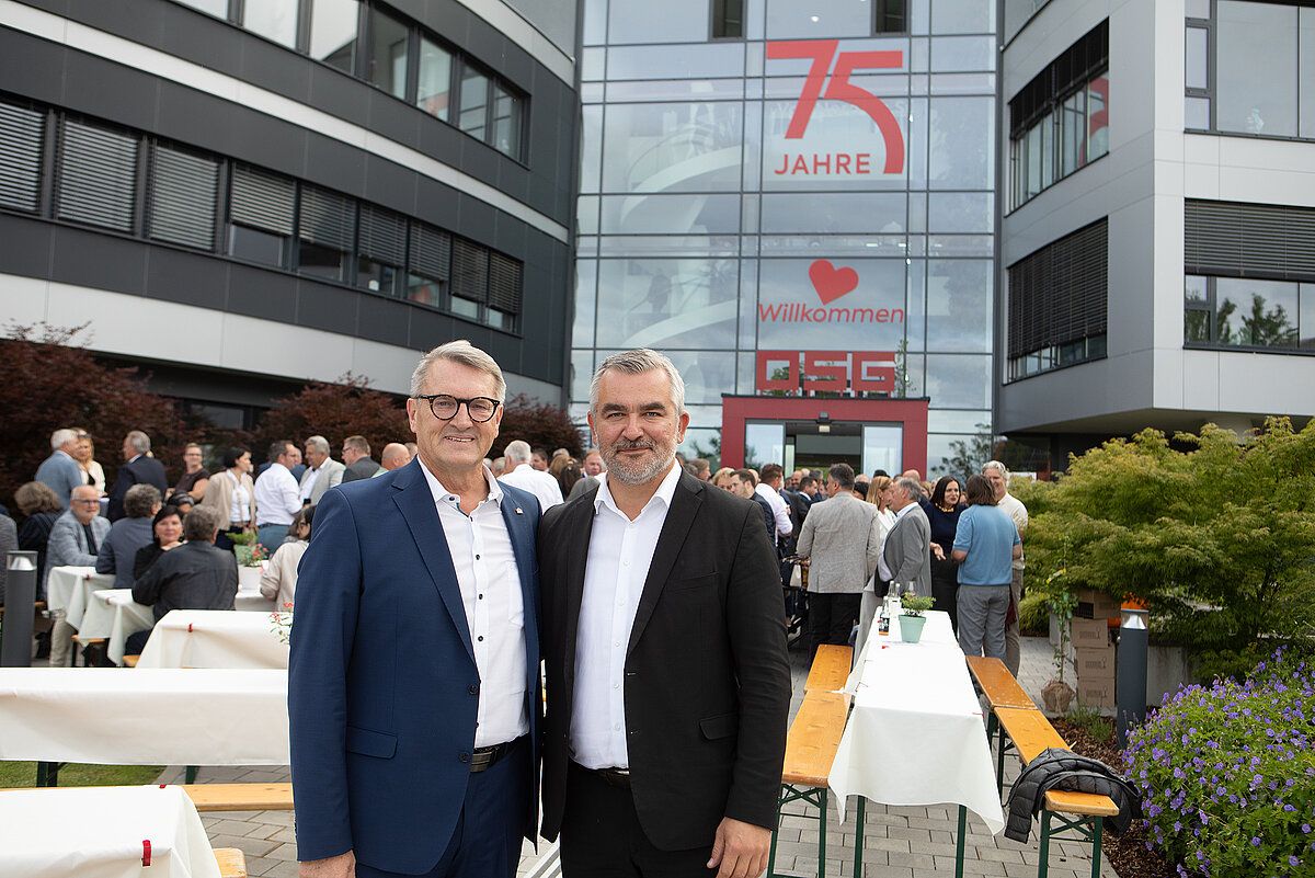 Two men stand in front of a building with a large sign that says 75 Jahre. They are smiling for a photo. Behind them is a crowd of people in a courtyard.