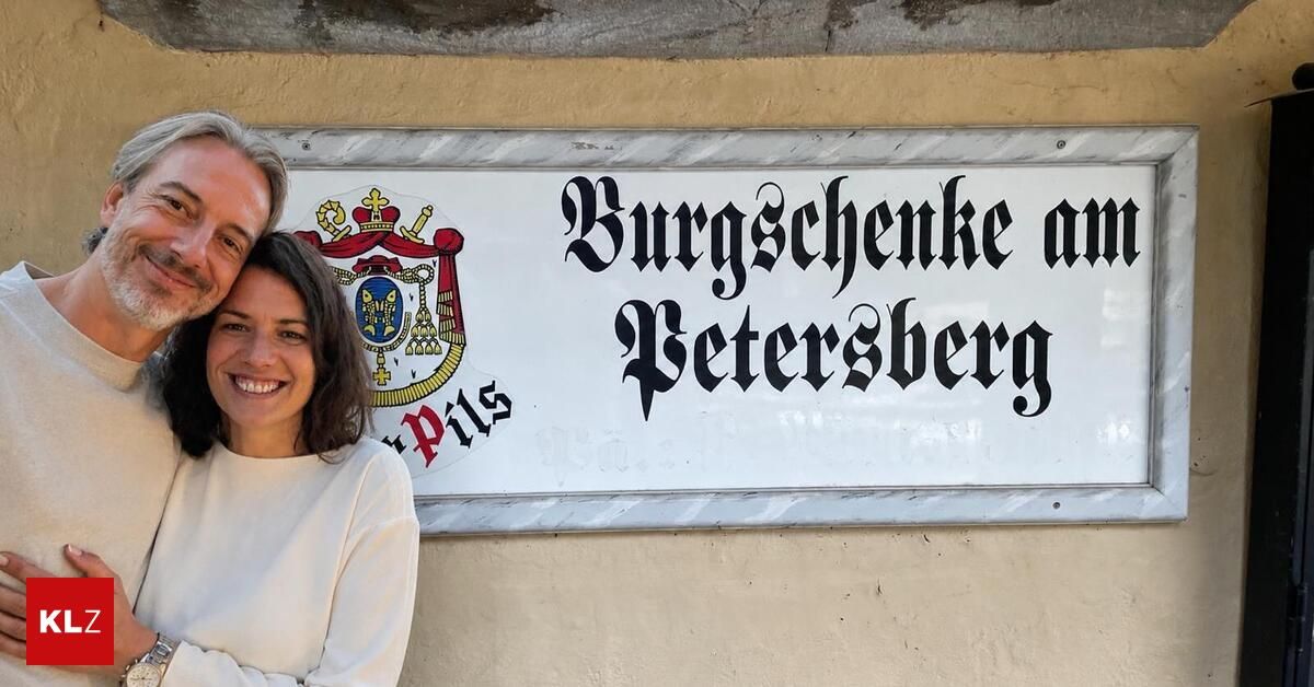 A smiling couple stands before a sign with the text 'Burgschenke am Petersberg', featuring a coat of arms and the word 'Pils'.