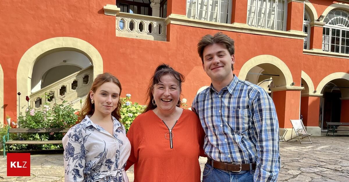 Three people stand outside an ornate orange building, smiling. A woman in the center wears a red shirt, while the man on the right wears a blue plaid shirt and a brown belt.