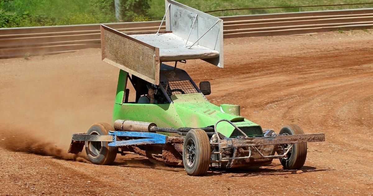 A green dirt racing car with a rusty roof drives on a dirt track, kicking up dust. A driver sits inside, gripping the steering wheel.
