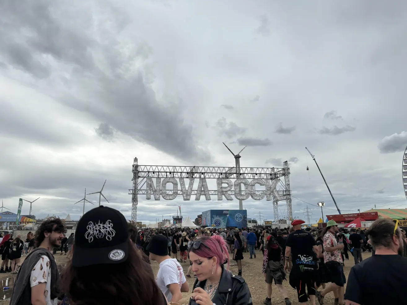 Eine Menschenmenge versammelt sich beim Nova Rock Festival unter einem bewölkten Himmel. Im Hintergrund steht ein großes Schild mit dem Festivalnamen.