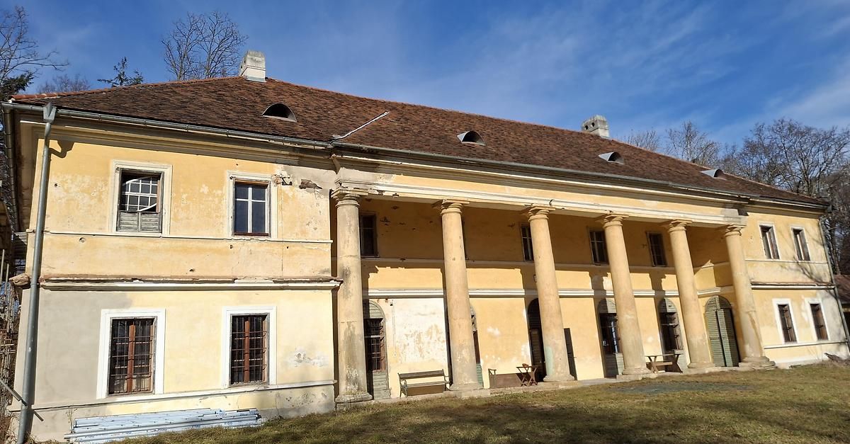 An old building with a brown roof and several pillars, benches, and a table outside. The building is painted yellow with windows, and it is surrounded by grass.