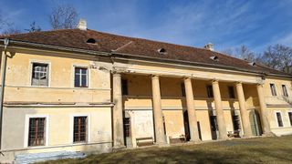 An old building with a brown roof and several pillars, benches, and a table outside. The building is painted yellow with windows, and it is surrounded by grass.