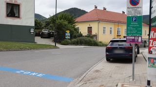 A yellow house with a red roof and several windows is on the side of the road. A blue car is parked on the curb.
