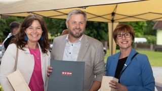 Three people stand under a tent, smiling. A man holds a certificate. A woman on the left wears a necklace and pink top. Another woman on the right wears glasses and holds a book.