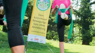 A woman exercises on a sunny day in a park, using a resistance band, standing near a yellow banner with text.