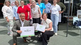 A group of people pose for a photo in front of a building. One holds a certificate, and others wear uniforms, indicating a healthcare setting.