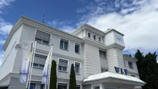A white building with 'WLW' on the entrance. There are two flags and trees in front. It is under a blue sky with clouds.