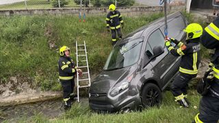 Three firefighters in yellow helmets are rescuing a car stuck in a river. A ladder and ropes are used to pull it out.