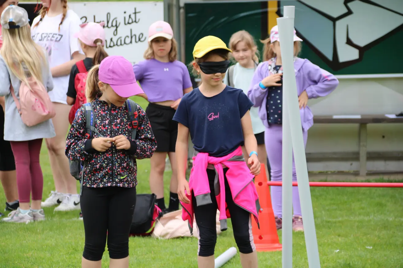 Eine Gruppe junger Mädchen nimmt an einer lustigen Aktivität im Freien teil. Eines der Mädchen trägt eine Augenbinde und einen rosa Schal, während die anderen Hüte tragen und zuschauen.