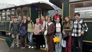 A group of people, both male and female, are standing in front of a train, smiling for a photo. They are wearing different colored jackets and holding bags. Behind them is a train with windows and a door.