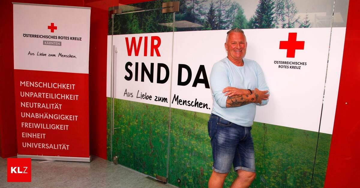 A smiling man stands in front of a banner that reads, 'We are here' with a green background. He wears a blue shirt and denim shorts.