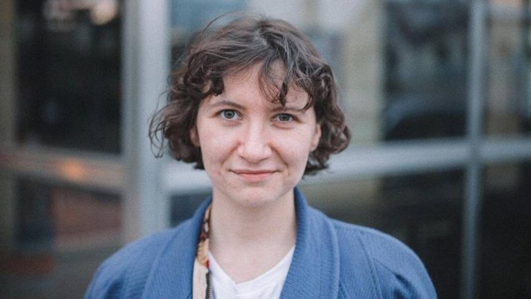 Eine lächelnde Frau mit lockigem Haar steht vor einem Gebäude. Sie trägt eine blaue Jacke und ein weißes Hemd. Der Hintergrund ist leicht unscharf.