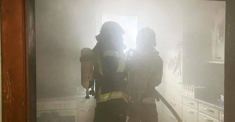 Zwei Feuerwehrleute in Schutzkleidung stehen in einer rauchigen Küche, einer hält einen Feuerlöscher und der andere einen Schlauch.