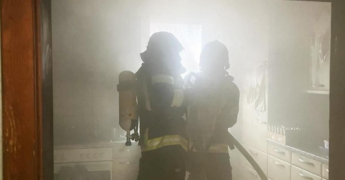 Two firefighters in protective gear stand in a smoky kitchen, one holding a fire extinguisher and the other with a hose.