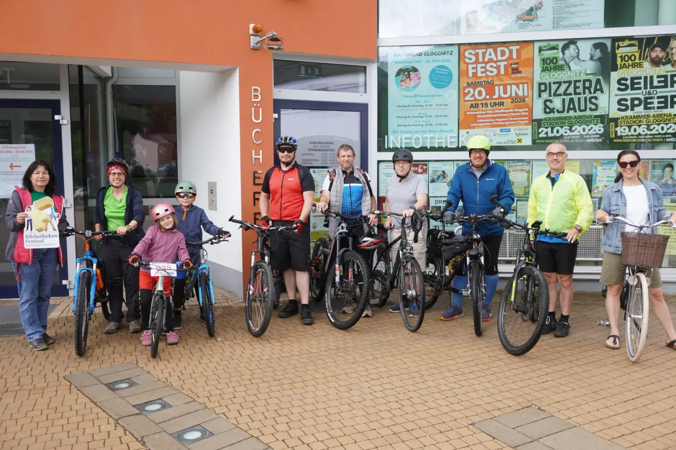 Eine Gruppe von Radfahrern, darunter Erwachsene und Kinder, posiert für ein Foto vor einem Geschäft mit einem Plakat zum Stadtfest im Hintergrund.