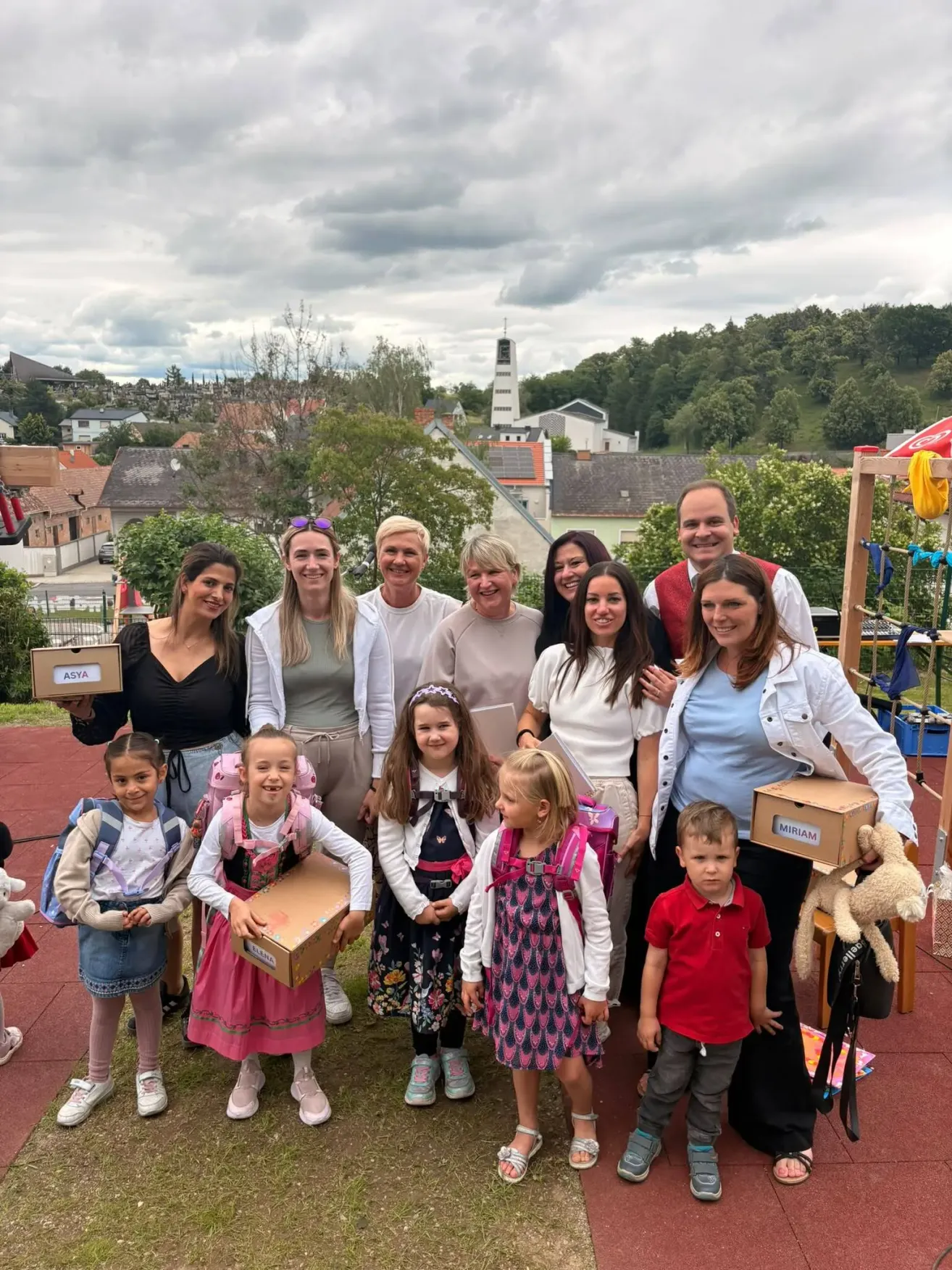 A group of people stand together in an outdoor setting. Adults and children smile for a photo. Some children hold boxes, and the background shows houses and trees.