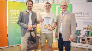 Three men stand in front of a green banner, one holding a certificate. The man in the middle wears glasses and a watch. On the left, a man carries a brown bag. The background has boxes and a white wall.