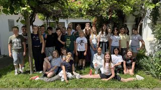 A group of children and teenagers are gathered outside, smiling and posing for a photo in front of a house with greenery.