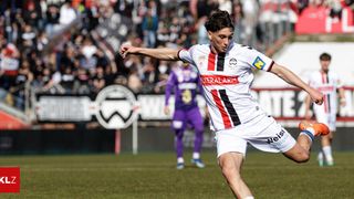 A soccer player wearing a white jersey with red and black stripes kicks the ball. Behind him, another player in purple is blurred, and spectators are watching.