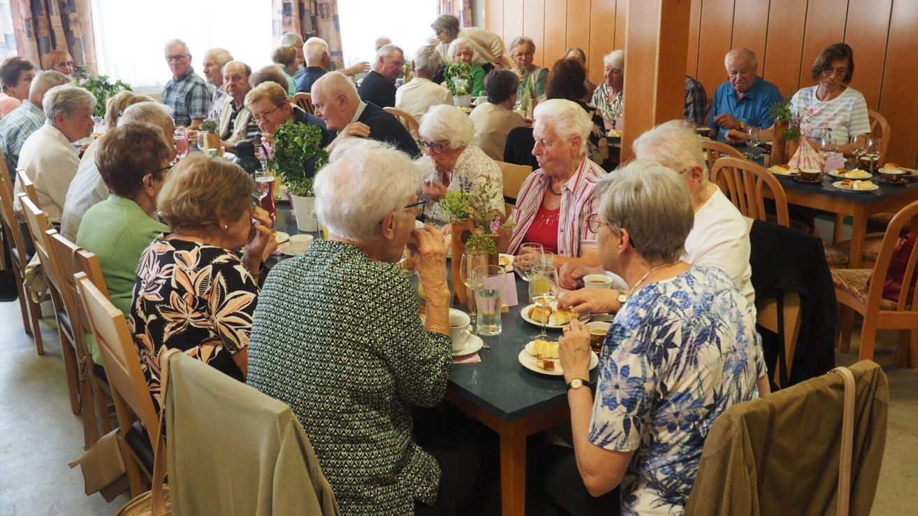 Elderly people sit at a long table, eating and drinking. They wear glasses and jewelry. A plant and cups are on the table. Behind them, people are sitting and standing in chairs.