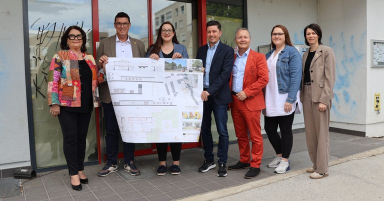 A group of people stands outside a building with a red door, holding a large architectural plan. They seem to be discussing the design.