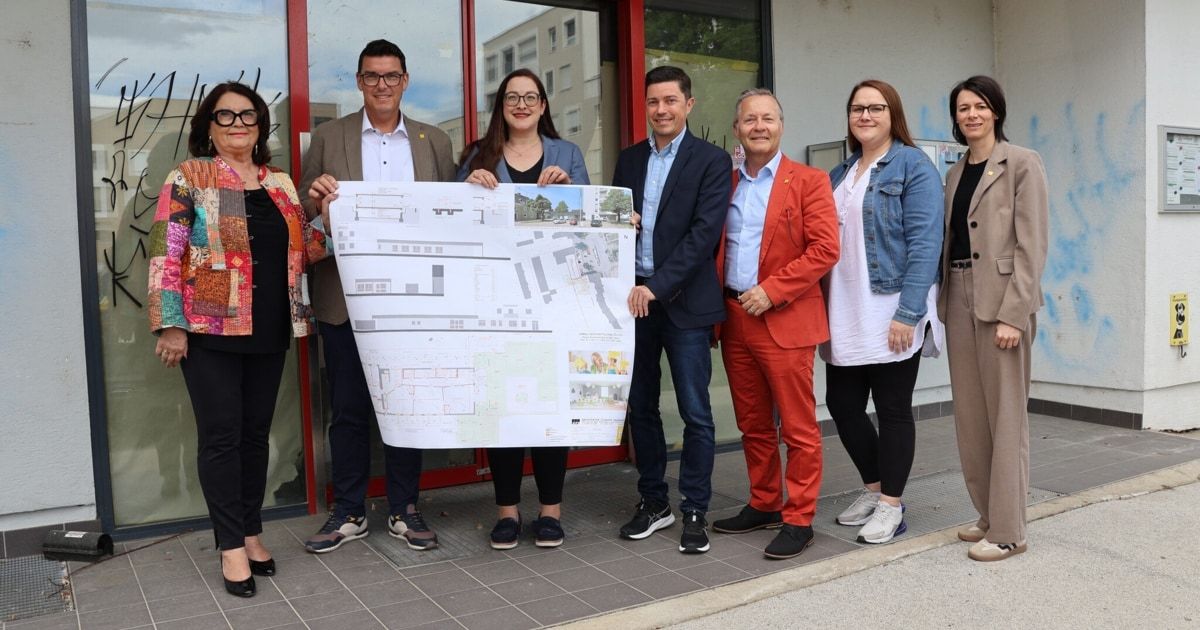 A group of people stands outside a building with a red door, holding a large architectural plan. They seem to be discussing the design.