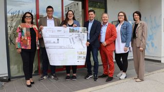 A group of people stands outside a building with a red door, holding a large architectural plan. They seem to be discussing the design.