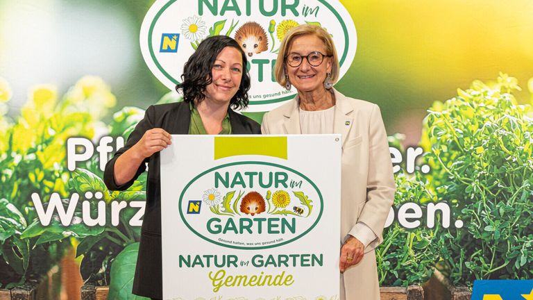 Zwei Frauen posieren mit einem Schild mit dem Titel 'Natur im Garten Gemeinde', das Blumen und ein Igel zeigt. Der Hintergrund ist grün mit einem Logo.
