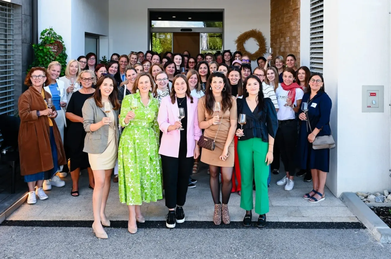 Eine Gruppe von Frauen in verschiedenen Kleidern, die lächeln und Weingläser halten, posiert für ein Foto vor einem Gebäude. Sie stehen auf einer Betonfläche mit einem dekorativen Kranz dahinter.