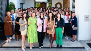 Eine Gruppe von Frauen in verschiedenen Kleidern, die lächeln und Weingläser halten, posiert für ein Foto vor einem Gebäude. Sie stehen auf einer Betonfläche mit einem dekorativen Kranz dahinter.
