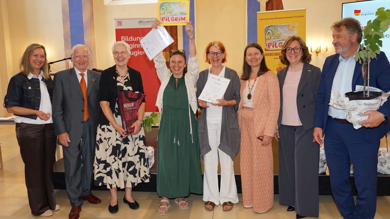 Eine Gruppe von Frauen steht lächelnd mit Urkunden und einem Buch. Eine Frau hält ein Schild mit der Aufschrift 'Pilger'. Im Hintergrund befinden sich Banner mit Text und Logos.